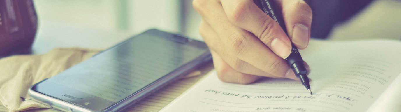 A person writing notes on a pad of paper with a phone lying next to them
