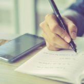 A person writing notes on a pad of paper with a phone lying next to them