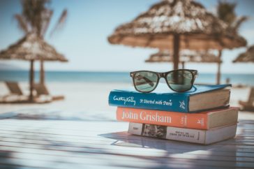 A stack of books by the oacean with sunglasses resting on top, and sun shades in the background